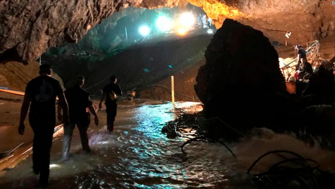 Rescue workers navigate through a partially flooded cave with equipment on the ground, surrounded by rock walls and illuminated by bright lights in the background.